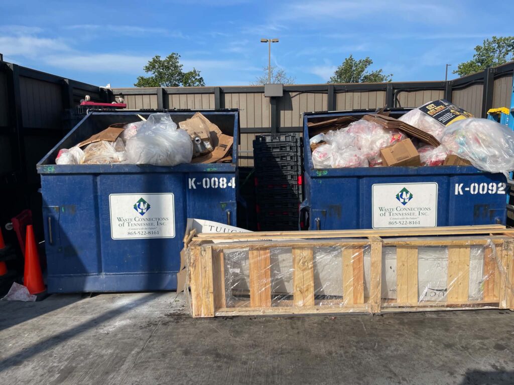 Blue commercial dumpsters with bags, cardboard and other material in the service area before cleanup.