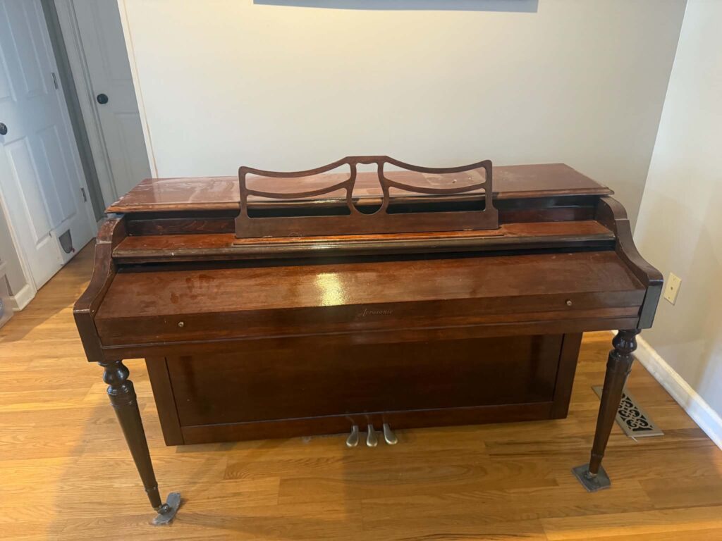 Wooden upright piano against a gray wall on hardwood flooring before removal.