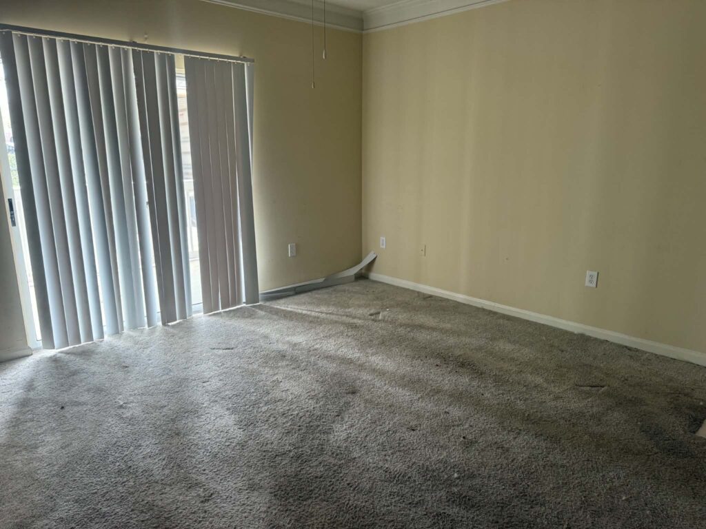 Empty carpeted apartment living room with vertical blinds and a sliding glass door after the furniture was removed