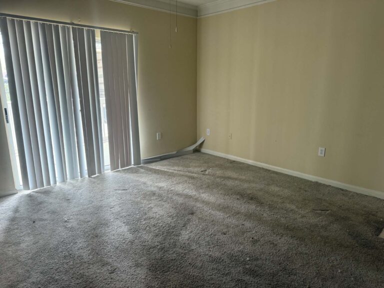 Empty carpeted apartment living room with vertical blinds and a sliding glass door after the furniture was removed