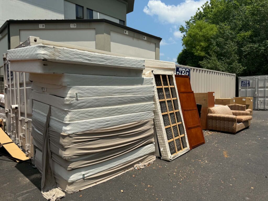 Mattresses and box springs stacked outside beside wooden doors and an upholstered chair during a bulk removal project.