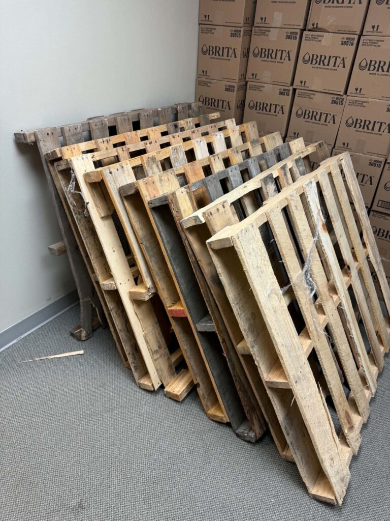 Wooden pallets in front of stacked delivery boxes inside an office storage area before pickup.