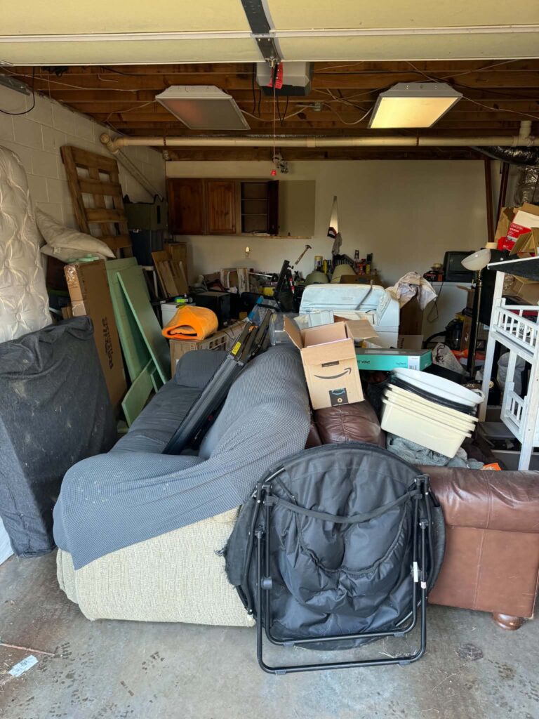 Upholstered furniture, boxes and loose items occupying the center of a garage with exposed ceiling rafters
