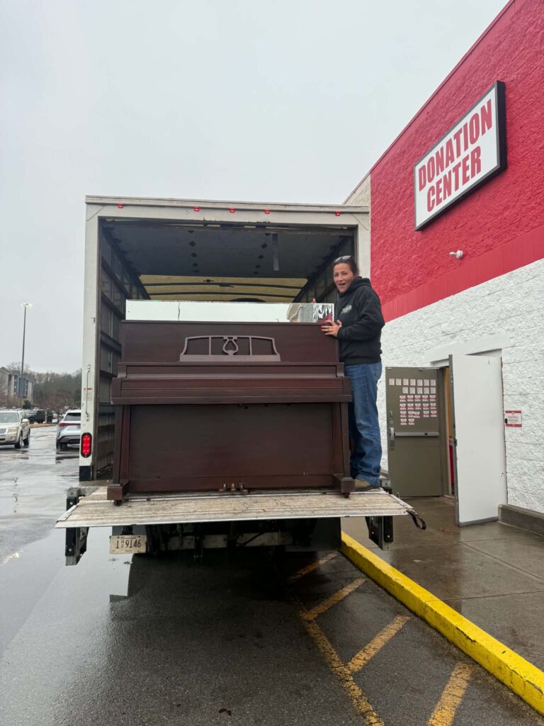 Crew member beside an upright piano on a truck at a donation center.