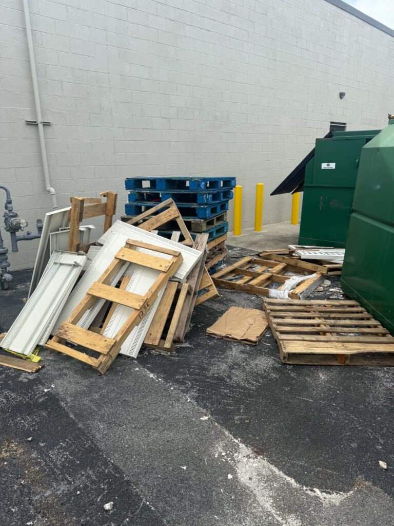 Wooden and blue pallets with metal shelving beside green dumpsters and a gray block wall before pickup at a commercial property.
