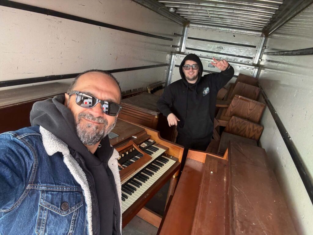 Two crew members inside a box truck with keyboard instruments and wooden church pews.