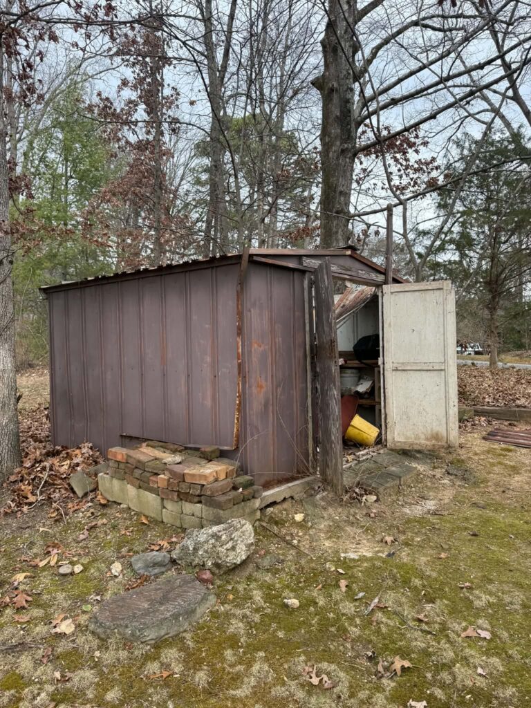 Small brown backyard shed with an open door and stored items inside, before removal.