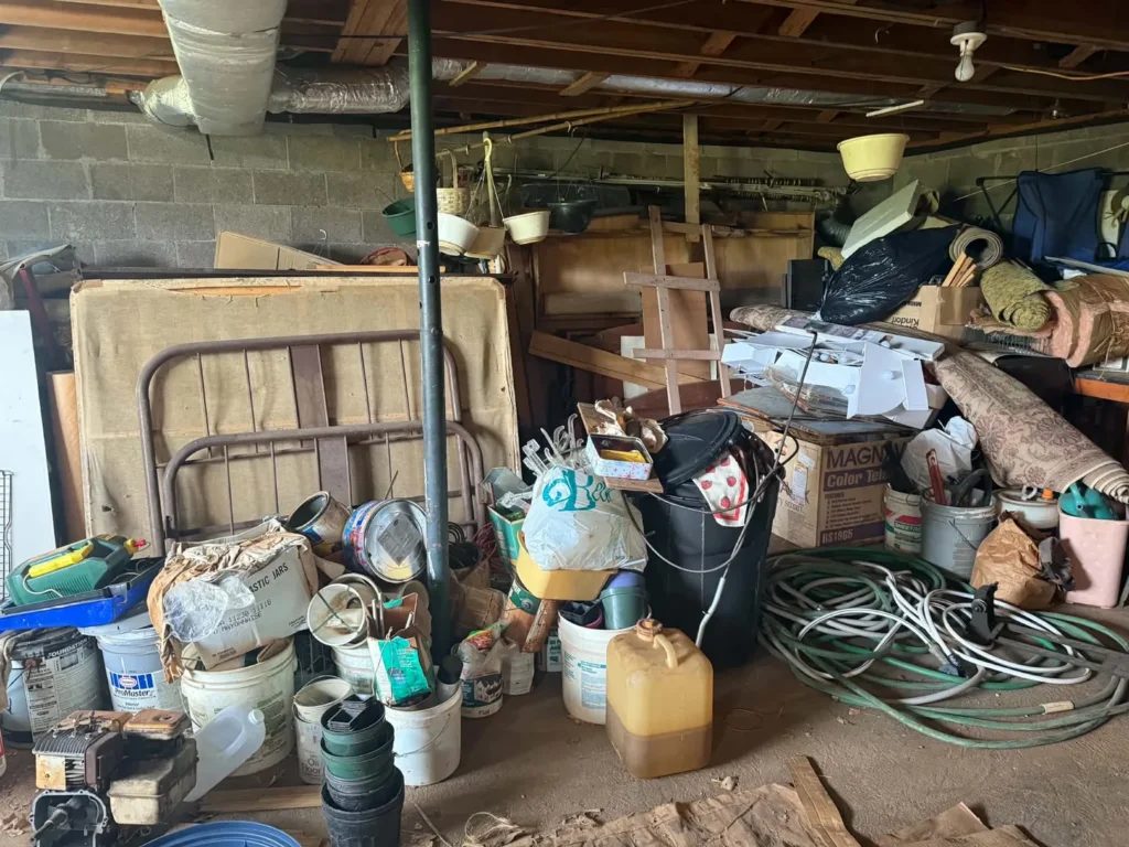 Furniture, rolled rugs, containers and other belongings occupying a basement floor.