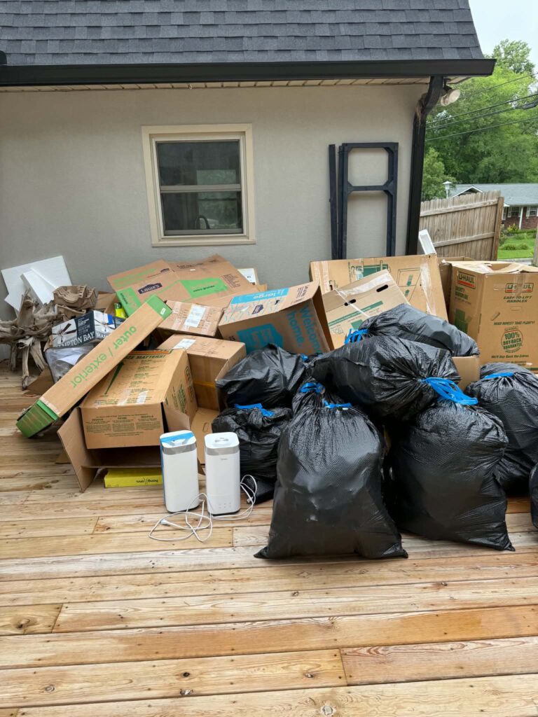 Cardboard moving boxes, packing materials, and bags piled on a wooden deck before pickup.
