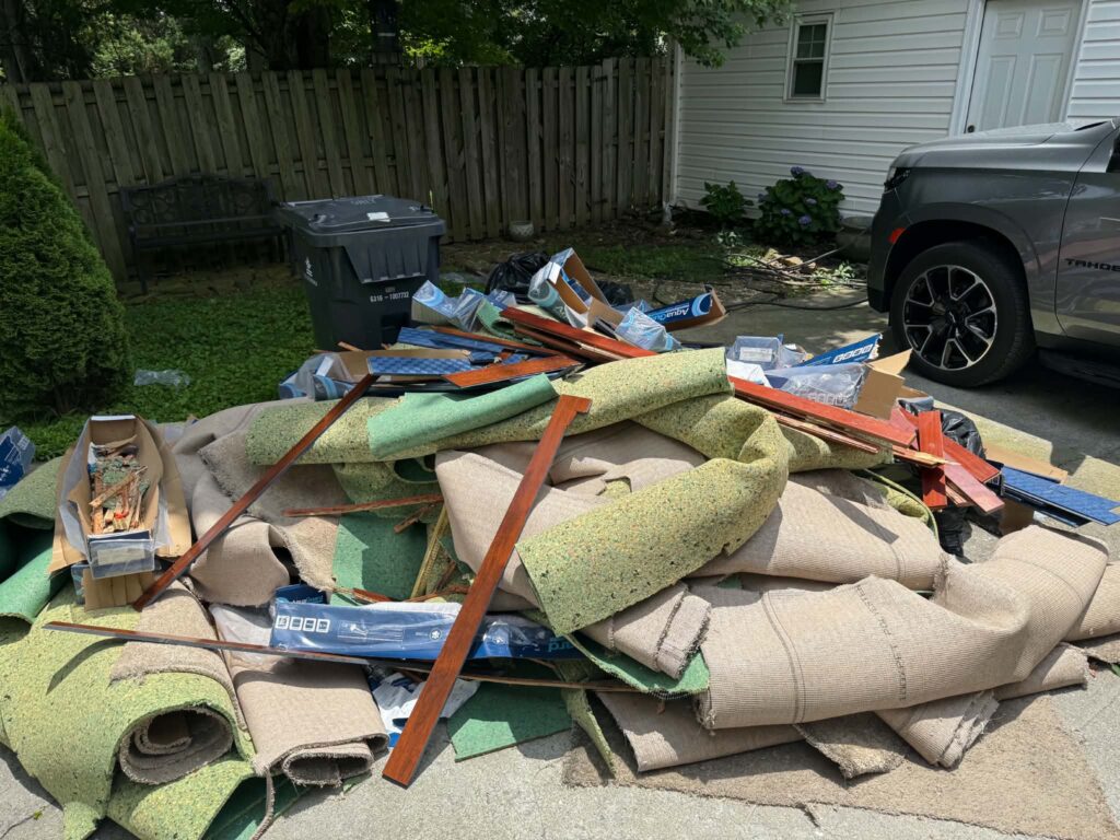Carpet rolls, padding, wood, and remodeling debris piled on a paved area before removal.